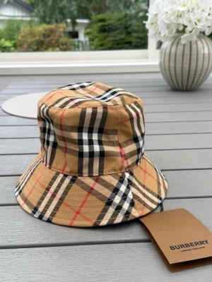 Burberry beige bucket hat - Säljer en klassisk bucket hat från Burberry i beige med det ikoniska rutmönstret i svart, vitt och rött. Hatten är tillverkad i bomull och har en avslappnad passform med bred skärm. Perfekt för dig som vill ha en trendig accessoar med lyxig känsla.
