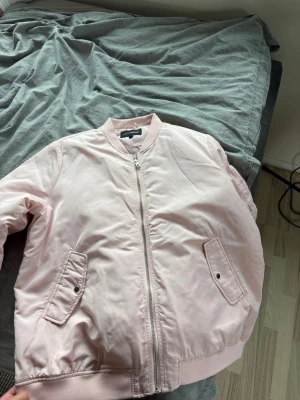Ljusrosa bomberjacka från Weekday 💗 - Snygg ljusrosa bomberjacka från Weekday med klassisk ribbad krage, dragkedja framtill och två fickor med knappstängning. Jackan har en stilren look och är tillverkad i ett glansigt material som ger en modern vibe. Perfekt för dig som vill sticka ut med en trendig jacka. Oanvänd 