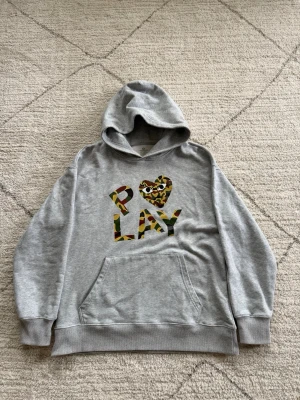 COMME DES GARÇONS PLAY hoodie - Snygg grå hoodie med stor ficka framtill och huva. På bröstet står det 'PLAY' i bokstäver med camouflage-mönster och ett hjärta med ögon. Perfekt för dig som gillar streetwear och vill sticka ut lite extra.