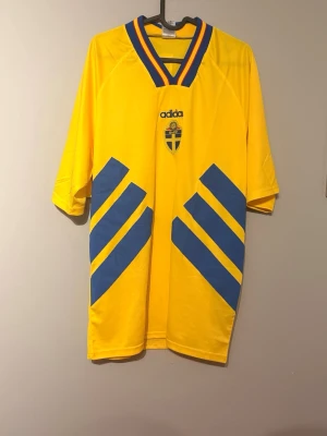 Gul Sverige fotbollströja Adidas - Säljer en klassisk gul fotbollströja från Adidas med blå detaljer och Sveriges landslagsemblem på bröstet. Tröjan har V-ringad krage med blå och gula ränder samt stora blå ränder på framsidan. Perfekt för match eller träning.