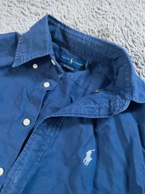Ralph Lauren skjorta  - Klassisk blå skjorta från Ralph Lauren med vit broderad logga på bröstet. Skjortan har lång ärm, knappar framtill och en avslappnad passform. Perfekt för en stilren och tidlös look.