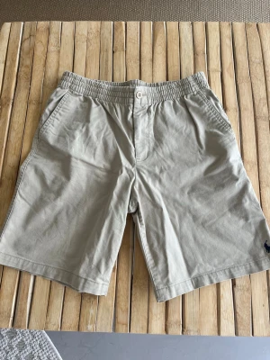Beige shorts från Polo Ralph Lauren - Klassiska beige shorts från Polo Ralph Lauren med elastisk midja och knappstängning. Tillverkade i mjuk bomull och har två sidofickor samt den ikoniska mörkblå loggan broderad vid benet. Perfekta för sommardagar och enkel att matcha.