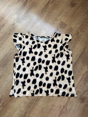 Beige topp med svart leopardmönster - Snygg topp från Lindex i beige med svart leopardmönster. Modellen har korta volangärmar och rund halsringning. Perfekt för dig som vill sticka ut lite extra med ett djurmönstrat plagg.