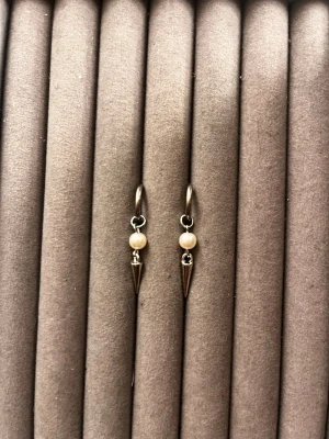 Silvriga örhängen med pärldetalj - Säljer ett par stilrena örhängen i silverfärg med rund vit pärldetalj och en spetsig metallhänge. Örhängena har en modern och edgy look med en liten ring som bas. Perfekta för dig som gillar minimalistiska accessoarer med en twist.