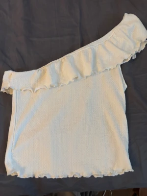 Vit oneshoulder topp med volang - Trendig vit oneshoulder topp med volangdetalj längs axeln och nedtill. Toppen är ärmlös och har en lätt strukturerad yta som ger en snygg touch. Perfekt för dig som vill sticka ut med en unik och stilren look.