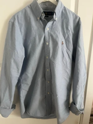 Ljusblå skjorta från Ralph Lauren - Klassisk ljusblå skjorta från Ralph Lauren med button-down krage och broderad logga på bröstet. Skjortan har långa ärmar, vita knappar och är tillverkad i mjuk bomull. Perfekt till jeans eller chinos för en clean och stilren look.