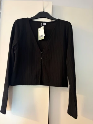 Svart ribbad kort kofta från H&M - Svart ribbad kort kofta från H&M med djup V-ringning och två knappar framtill. Långärmad och i ett mjukt, stretchigt material som sitter snyggt på kroppen. Perfekt att styla med jeans eller kjol för en trendig look.