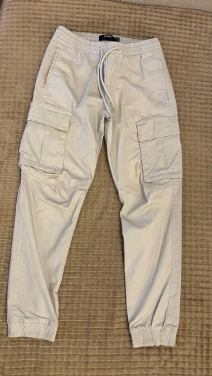 Beige cargobyxor från Pull & Bear - Säljer ett par snygga beige cargobyxor från Bull & Bear med resår i midjan och benslut. Byxorna har stora sidofickor med lock, dragsko i midjan och en avslappnad passform. Perfekta för en chill och trendig look.