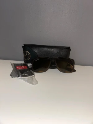 Svarta Ray-Ban solglasögon - Snygga svarta solglasögon från Ray-Ban med rektangulär form och mörka glas. Klassisk design med Ray-Ban-logga på linsen. Kommer med originalfodral och putsduk. Perfekta för soliga dagar och ger en cool vibe till din outfit.