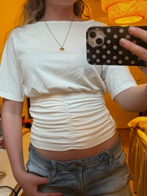 Vit croppad topp med rynkdetalj - Snygg vit croppad topp med rynkade detaljer över magen och korta ärmar. T-shirten har en enkel, stilren design och är perfekt att matcha med jeans eller kjol. Materialet känns mjukt och stretchigt, vilket gör den bekväm att bära.