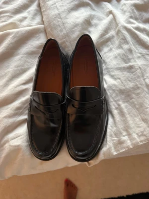 Svarta loafers  - Säljer ett par stilrena svarta loafers i blankt skinn med klassisk penny-loafer design. Skorna har rundad tå och diskret söm längs kanten. Insidan är fodrad i brunt läder för extra komfort. Perfekta för en clean och tidlös look.