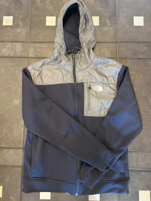Blå hoodie från The North Face - Snygg blå hoodie från The North Face med dragkedja och grå detaljer på axlar och huva. Bröstficka med dragkedja och logga på vänster sida. Tillverkad i mjukt fleecefoder och slitstarkt material, perfekt för chill eller utomhusaktiviteter.