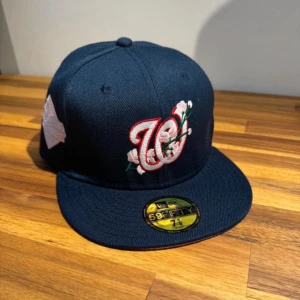 Mörkblå New Era keps med brodyr - Snygg mörkblå keps från New Era 59FIFTY med broderad röd och vit W-logga och blommor framtill. Platt skärm och klassisk passform. Perfekt för dig som vill sticka ut med en unik detalj. Märke och storlek syns på klistermärket på skärmen.