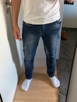 Blå slim fit jeans med slitningar från Sams - Säljer ett par blå slim fit jeans med lätt slitna detaljer på låren och knäna. Jeansen har klassisk femficksdesign och normal midja. Materialet är denim med en skön stretch som ger bra passform. Perfekta för en avslappnad och trendig look.
