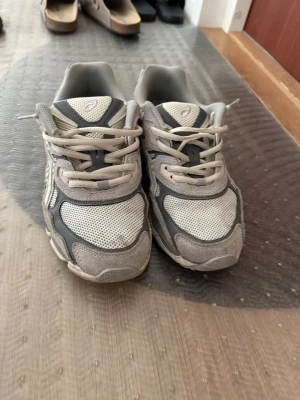 Gråa Asics sneakers med meshdetaljer - Snygga sneakers från Asics i grått och vitt med klassisk logga på sidan. Skorna har meshpaneler för extra andningsförmåga och detaljer i mocka och syntet. Perfekta för en sportig och avslappnad stil. Lågt skaft och snörning framtill.