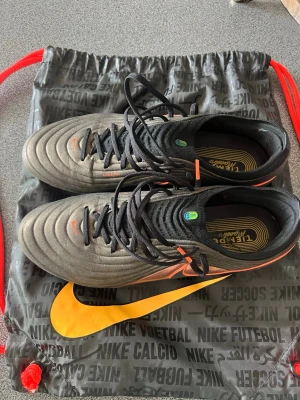 Nike Tiempo maestro elite fotbollsskor - Snygga Nike Tiempo maestro elite fotbollsskor i svart och koppar med orange detaljer. Skorna har klassisk snörning, mönstrad ovandel i syntetmaterial och FG-dobbar för spel på naturgräs. Perfekta för dig som vill ha bra touch och stabilitet på planen. Använda tre gånger eftersom de var lite för små.