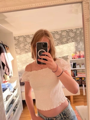 Vit offshoulder blus med spetsdetaljer - Supersöt vit topp med smockad kropp och korta puffärmar i broderad spets. Toppen har offshoulder-modell och volangdetaljer vid ärmslut och nederkant. Perfekt för en fräsch och somrig look. Den är storlek L men jag har S och den passar mig bra
