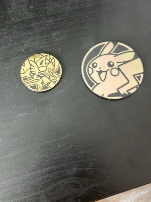 Pokémon mynt med Pikachu och Zamazenta - Två Pokémon mynt i plast med motiv av Pikachu och Zamazenta. Ena myntet är större med Pikachu och det andra mindre med Zamazenta, båda i guld och svart design. Perfekt till Pokémonkortspel eller samling.