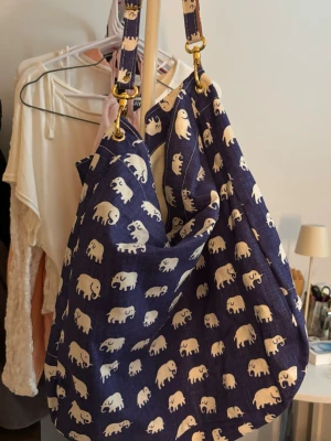 Blå shoppingväska med elefanter - Snygg väska från Svenskt Tenn i blått tyg med vitt elefantmönster. Väskan har bred axelrem och guldfärgade metalldetaljer. Pris kan diskuteras💗