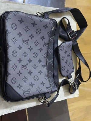  axelremsväska - Snygg axelremsväska i svart och grått med klassiskt monogrammönster. Väskan har justerbar axelrem, silverfärgade detaljer och en matchande liten pouch. Tillverkad i tåligt canvasmaterial med läderdetaljer och dragkedja upptill.