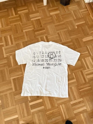 Maison Margiela vit t-shirt  - Vit t-shirt från Maison Margiela med svart tryck på ryggen som visar märkets ikoniska siffror och logga. Klassisk passform med korta ärmar och rund hals. Tillverkad i mjuk bomull, perfekt för en clean och trendig look.