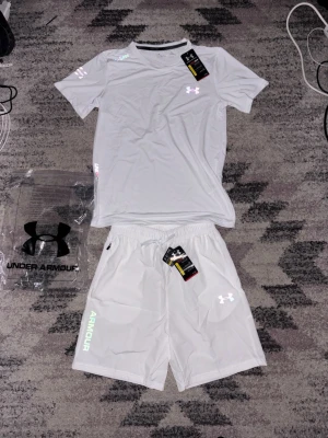 Vitt träningsset från Under Armour - Snyggt vitt träningsset från Under Armour med t-shirt och shorts. Setet har diskreta loggor och detaljer i reflekterande material. Tillverkat i lätt och snabbtorkande funktionsmaterial som passar perfekt till gymmet eller löprundan.