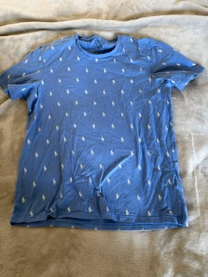 Blå Polo Ralph Lauren t-shirt - Snygg blå t-shirt från Polo Ralph Lauren med klassiskt crewneck och små vita polospelare tryckta över hela tröjan. Tillverkad i mjuk bomull som känns skön mot huden. Perfekt för en avslappnad och stilren look.