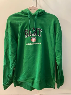 Grön hoodie från GANT - Grön hoodie från GANT med klassiskt tryck på bröstet och texten 'AMERICAN SPORTSWEAR'. Hoodien har huva med dragsko och ribbade muddar vid ärmslut och nederkant. Använd 2-3 gånger. Litet hål (knappt märkbart) bak på luvan (se bild). Nypris 1300kr