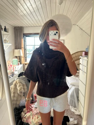 Brun stickad poncho med polokrage - Säljer en brun stickad poncho med bred polokrage och korta ärmar. Ponchon har ett snyggt flätmönster framtill och bred ribbstickad kant nedtill. Perfekt att slänga över en tröja för extra värme och stil.