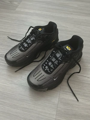 Nike Air Max Plus TN Svart/Grå - Nike Air Max Plus TN sneakers i svart och grå med coola detaljer och synlig Tn-logga på plösen och sidan. Skorna har en sportig vibe med svarta snören, syntetiskt material och distinkta linjer på ovandelen. Perfekta för dig som gillar streetwear och snygg design.