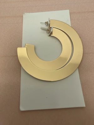 Stora guldiga cirkelörhängen - Säljer ett par stora, chunky örhängen i guldig metall med modern, öppen cirkelform. De har en slät och blank yta som ger en cool statement-look. Perfekta för dig som gillar att sticka ut med accessoarer.