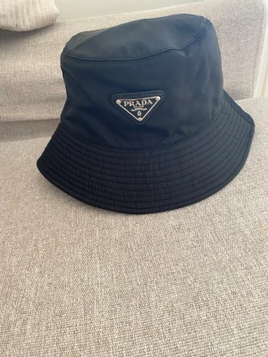 Svart bucket hat från Prada - Snygg svart bucket hat från Prada i nylon med det klassiska trekantiga metalloggan framtill. Hatten har bred skärm och stilren design som passar perfekt till streetwear och trendiga outfits.