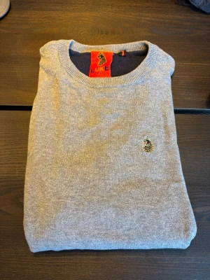 Grå stickad tröja från LUKE - Snygg grå stickad tröja från LUKE med rund halsringning och diskret broderad logga på bröstet. Tröjan har ribbade muddar vid ärmslut och nederkant, perfekt för en clean och stilren look. Pris kan diskuteras!