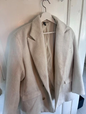 Vit ullkappa från H&M - Snygg vit kappa från H&M  med bred krage och två fickor framtill. Kappan har rak passform och stängs med knappar. Perfekt för dig som gillar en clean och modern look till höst och vinter.