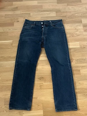 Mörkblå raka Levi's jeans - Klassiska mörkblå jeans från Levi's med rak passform och knappgylf. Jeansen har fem fickor och en brun läderpatch bak i midjan. Tillverkade i slitstark denim som ger en tidlös look. Perfekta för dig som gillar en enkel och clean stil. Dom är lagade i grenen.