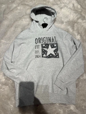 Grå hoodie med stjärntryck från SCNDI - Snygg grå hoodie med huva och trycket 'ORIGINAL FIT EST. 2024' samt en stor vit stjärna och små stjärnor på bröstet. Klassisk känguruficka framtill och långa ärmar. Perfekt för en avslappnad och trendig look.