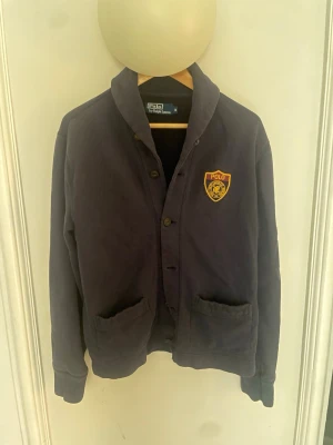 Mörkblå cardigan från Polo Ralph Lauren - En stilren Mörkblå cardigan med knäppning framtill från Polo Ralph Lauren. Tröjan har två stora fickor och ett broderat Polo-märke på bröstet i gult och vinrött. Skön och avslappnad passform med ribbade muddar vid ärmslut och nederkant.