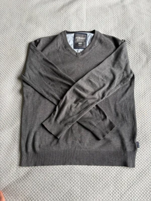 Grå V-ringad tröja från Sebago - Klassisk grå V-ringad tröja från Sebago Marine Classics. Tillverkad i mjukt material med ribbade muddar vid ärmslut och nederkant. Diskret logga broderad på bröstet. Perfekt att slänga på sig över en skjorta för en clean look. Det är dock en liten skada i höger armhåla som man ser på bild nummer 4. 