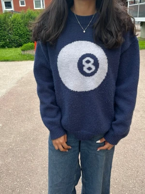 Mörkblå stickad tröja med 8-ball - Säljer en mörkblå stickad tröja med en stor vit 8-ball på bröstet. Tröjan har avslappnad passform, ribbade muddar och rund hals. Perfekt för dig som gillar streetstyle och vill ha något unikt i garderoben.