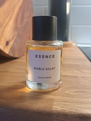 ESSNCE Noble Eclat Eau de Parfum - ESSNCE Noble Eclat Eau de Parfum i en stilren, rund glasflaska med svart lock. Doften har en elegant och minimalistisk design med vit etikett och svart text. Perfekt för dig som gillar exklusiva och moderna parfymer.