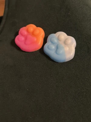Två tassformade squeshi - Säljer två suddgummin formade som tassar, en i orange och rosa samt en i blått och vitt. Perfekta att ha i pennfodralet eller som dekoration på skrivbordet. Gjorda av mjukt gummimaterial och har en söt design.
