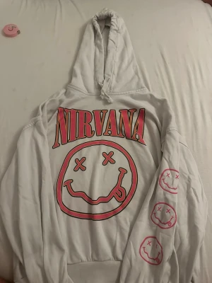 Vit Nirvana hoodie med rosa tryck - Säljer en vit hoodie med huva från Nirvana. Stor rosa logga och smiley-ansikte på bröstet samt tre mindre smileys på ena ärmen. Hoodien har dragsko i huvan och ribbade muddar. Perfekt för dig som gillar bandmerch och streetwear.