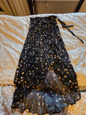 Svart blommig omlottkjol - Svart långkjol med små blommor i gult och rosa. Kjolen har omlottmodell med knytband i midjan plus knappar för extra support.. Tunt, luftigt material som passar perfekt till varmare dagar. Snygg och trendig för dig som gillar romantisk stil.