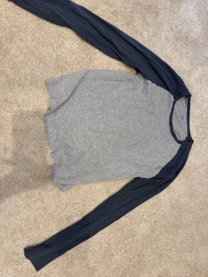 Grå långärmad topp från Brandy Melville  - En enkel och stilren långärmad topp med grå kropp och marinblå ärmar. Toppen har rund halsringning och är tillverkad i mjuk bomull, perfekt för en avslappnad look.