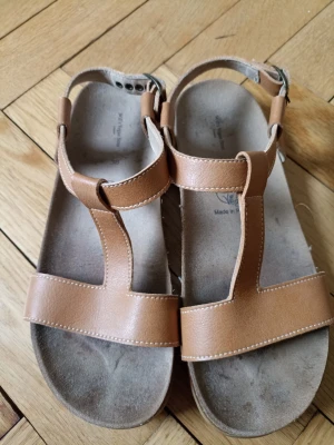 Bruna T-rem sandaler i läderlook - Snygga sandaler i klassisk T-rem design med justerbart spänne runt ankeln. Tillverkade i ljusbrun läderimitation med vita sömmar och mjuk innersula för extra komfort. Perfekta för sommardagar och passar till många olika outfits.