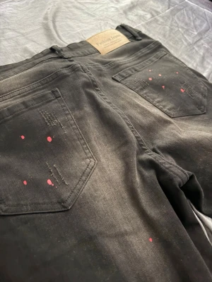 Svarta jeans med färgstänk och slitningar - Säljer ett par svarta jeans från Fashion Jeans med coola röda och vita färgstänk, slitna detaljer och patch med paljetter på ena benet. Jeansen har raka ben och två bakfickor. Perfekta för dig som gillar en edgy och unik stil.