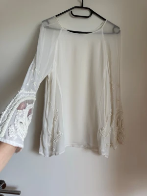 Vit transparent blus med pärlbroderi - Säljer en elegant vit blus i transparent mesh med långa ärmar. Ärmarna har detaljerade pärlbroderier och utskurna partier som ger en unik look. Perfekt för dig som vill sticka ut med något extra.