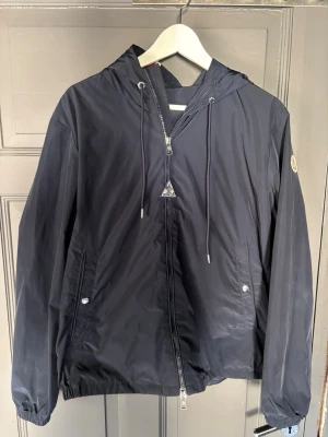 Mörkblå vindjacka från Moncler - Snygg mörkblå vindjacka från Moncler med huva. Jackan har två fickor med tryckknappar, dragkedja framtill och Moncler-logga på ärmen. Materialet är tunt och vattenavvisande, perfekt för blåsiga dagar. Detaljer i silver och diskret rödvit rand på huvan.