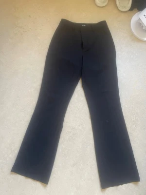 Svarta utsvängda kostymbyxor - Säljer ett par svarta kostymbyxor med utsvängda ben och hög midja. Klassisk design med diskreta fickor och knappstängning fram. Perfekta för en stilren och trendig look. Materialet känns mjukt och följsamt. Oanvänd