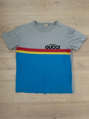 Flerfärgad Gucci t-shirt med tryck - Säljer en snygg t-shirt från Gucci med grå överdel, blå nederdel och färgglada ränder i rött och gult över bröstet. T-shirten har korta ärmar och ett tryck med texten 'Original Gucci' på bröstet. Tillverkad i mjuk bomull och perfekt för dig som gillar streetwear.